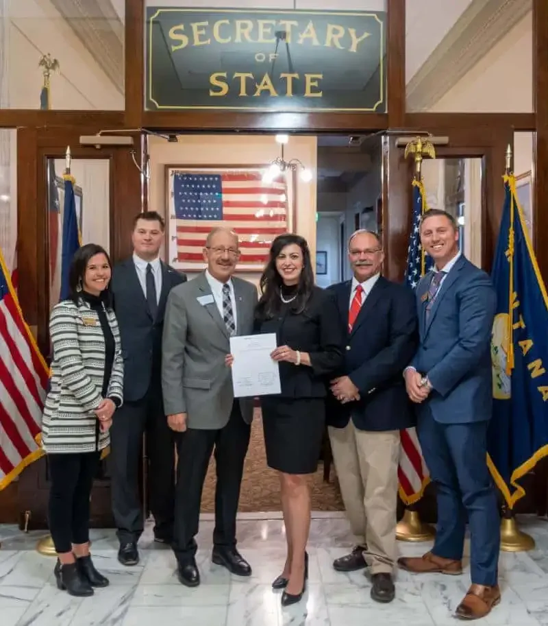 Six people stand smiling in front of a Secretary of State sign, American flag, and state flags inside a government building. The woman in the center holds a document, marking annual maintenance of voter rolls with Secretary Jacobsen.