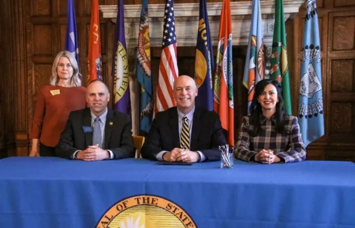 Secretary Jacobsen's priority bill revising election laws related to minor parties signed to Montana law 2 Four people pose at a table in front of state flags, likely discussing Montana election laws. Three are seated and one stands behind them. The blue-clothed table bears a seal, with pens arranged before the person in the center.