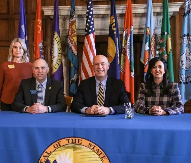 Four people pose at a table in front of state flags, likely discussing Montana election laws. Three are seated and one stands behind them. The blue-clothed table bears a seal, with pens arranged before the person in the center.