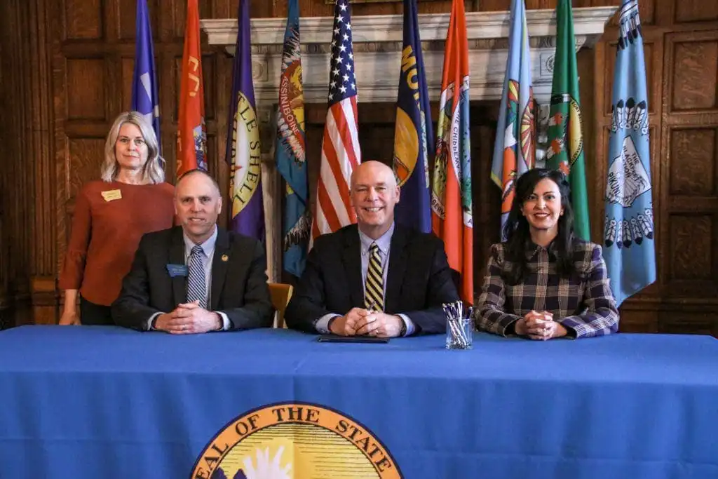 Four people pose at a table in front of state flags, likely discussing Montana election laws. Three are seated and one stands behind them. The blue-clothed table bears a seal, with pens arranged before the person in the center.