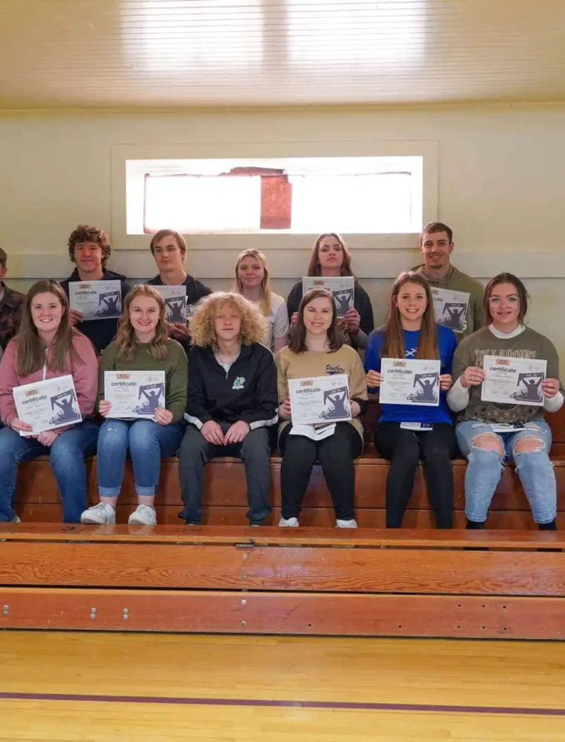 A group of students and two adults sit on wooden bleachers indoors at Sheridan High School. Many of the students, holding certificates from the US Constitution Contest, smile at the camera as light enters through a window behind them.