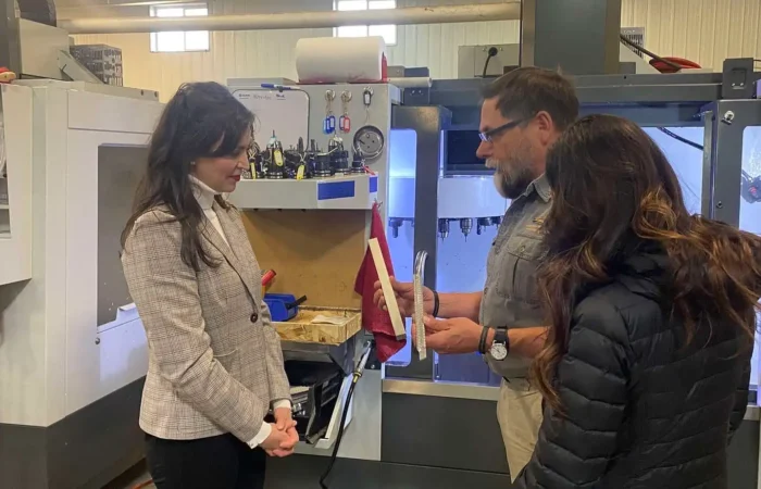 Montana Business Spotlight: Montana-made Skinner Sights trusted around the world 10 Three people stand in a workshop by industrial machinery. One man explains or shows a document to two women; Skinner Sights tools and equipment, known for being Montana-made, are visible in the background as the group appears engaged in discussion.