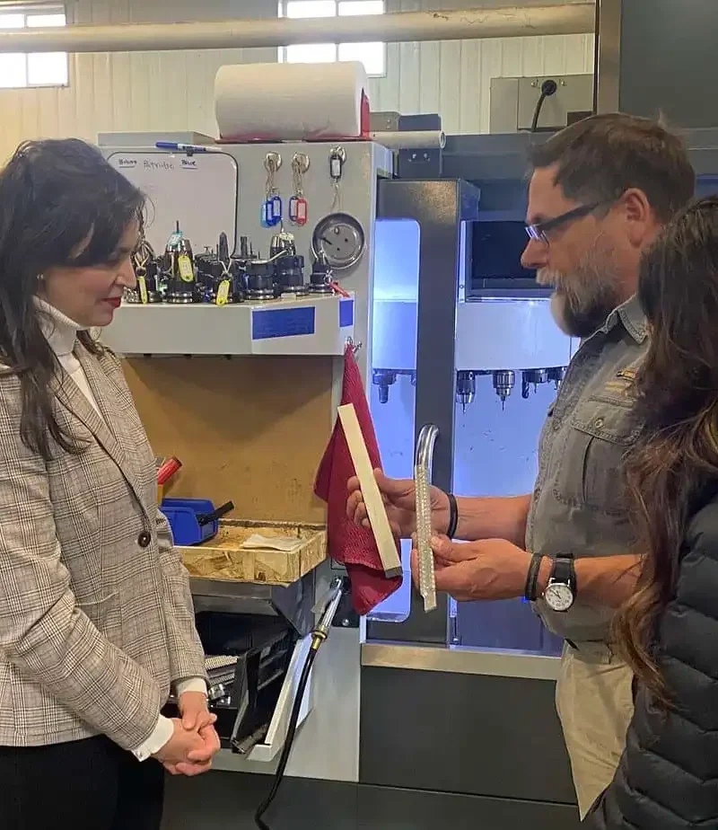 Three people stand in a workshop by industrial machinery. One man explains or shows a document to two women; Skinner Sights tools and equipment, known for being Montana-made, are visible in the background as the group appears engaged in discussion.