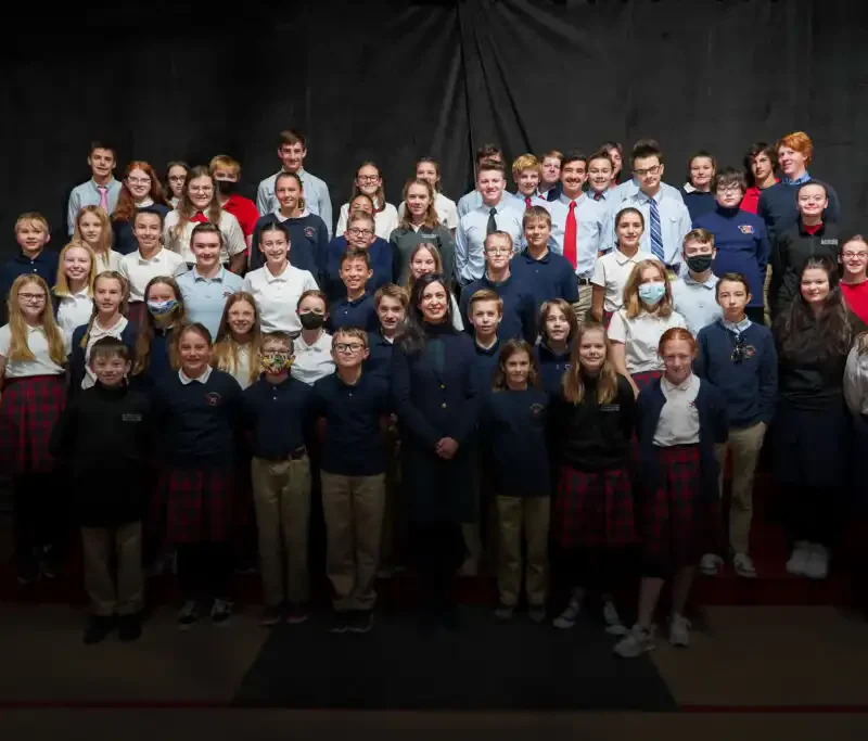 A large group of St. Andrew School middle school students in uniforms pose for a class photo in rows against a dark backdrop, with some students smiling and others looking serious, as Secretary Jacobsen observes nearby.