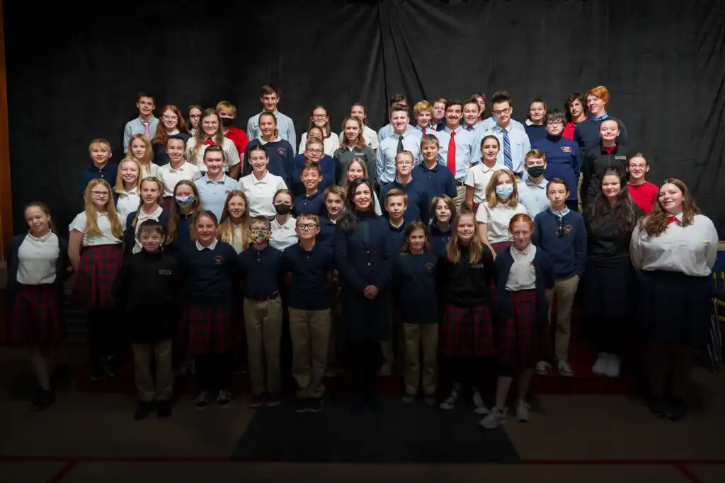 A large group of St. Andrew School middle school students in uniforms pose for a class photo in rows against a dark backdrop, with some students smiling and others looking serious, as Secretary Jacobsen observes nearby.