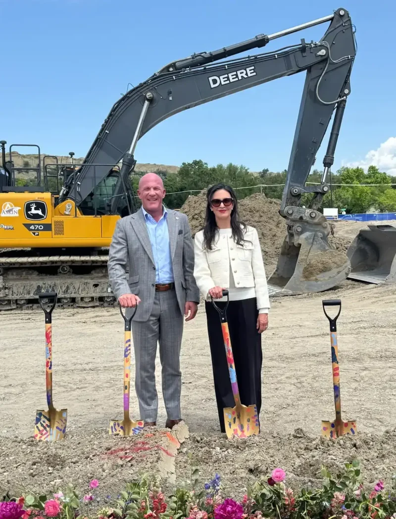 Christi Jacobsen stands smiling with a companion in front of colorful shovels at the St. Vincent Regional Hospital groundbreaking, with a yellow excavator, bulldozer, and bright pink flowers completing the scene.