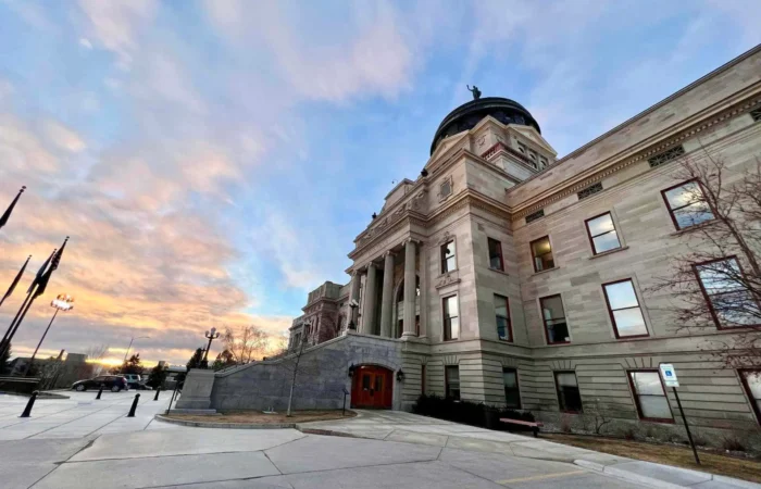 Secretary Christi Jacobsen shares September's new business registration update 9 The photo shows a grand stone building with columns and a large dome under a colorful sunset sky, capturing the spirit of Montana cities. Flags and trees add charm, with a wheelchair parking spot visible in the foreground.