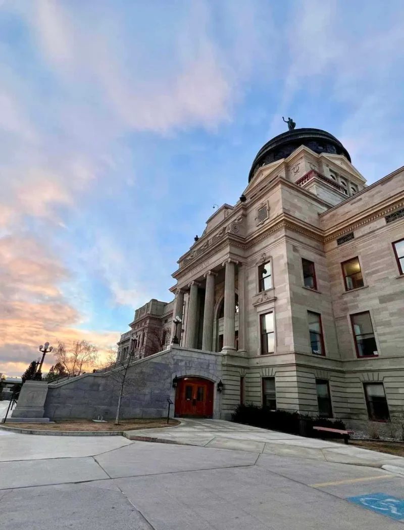 Four Montana cities on WalletHub's '2022 Best & Worst Small Cities to Start a Business' list 6 The photo shows a grand stone building with columns and a large dome under a colorful sunset sky, capturing the spirit of Montana cities. Flags and trees add charm, with a wheelchair parking spot visible in the foreground.