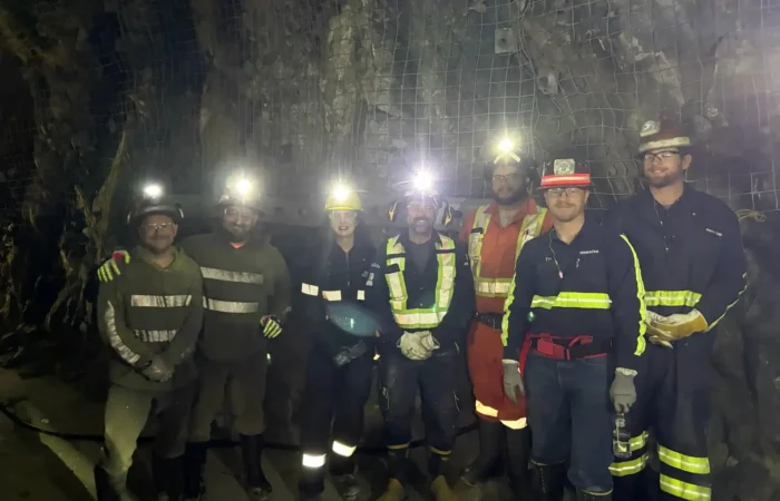 Exploring Montana's mining heritage: Secretary Christi Jacobsen commends Stillwater Mine team following recent visit 4 A group of seven workers wearing helmets with headlamps and reflective safety gear stand together in an underground tunnel with rocky walls and wire mesh at the Stillwater Mine, a cornerstone of Montana mining.