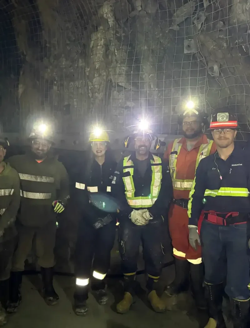 A group of seven workers wearing helmets with headlamps and reflective safety gear stand together in an underground tunnel with rocky walls and wire mesh at the Stillwater Mine, a cornerstone of Montana mining.