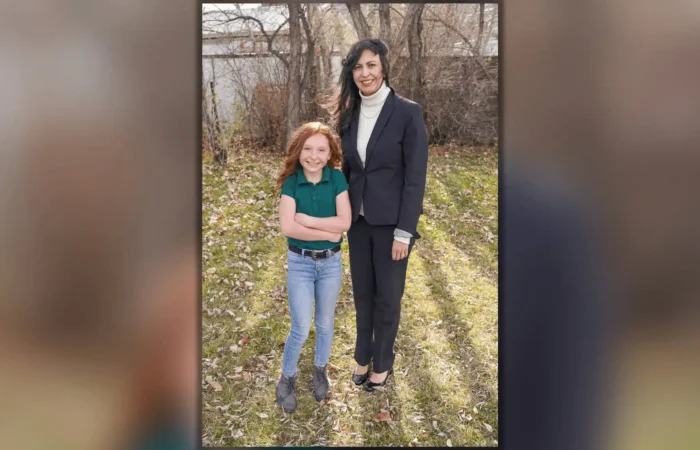 Song from The Way Christian School student wins US Constitution Contest 2 A woman in a dark suit stands outside on grass next to a smiling young girl with long red hair, wearing a green shirt and blue jeans. They appear to be celebrating the US Constitution Contest at The Way Christian School, with trees and a wooden fence behind them.