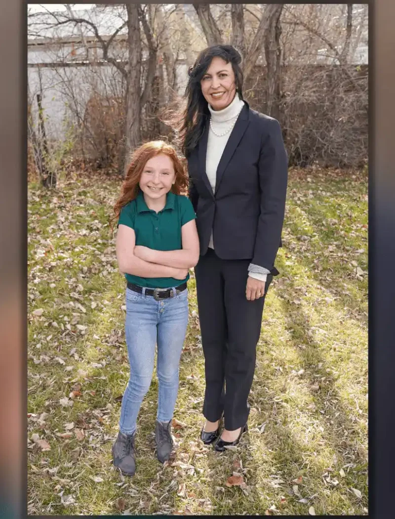 A woman in a dark suit stands outside on grass next to a smiling young girl with long red hair, wearing a green shirt and blue jeans. They appear to be celebrating the US Constitution Contest at The Way Christian School, with trees and a wooden fence behind them.