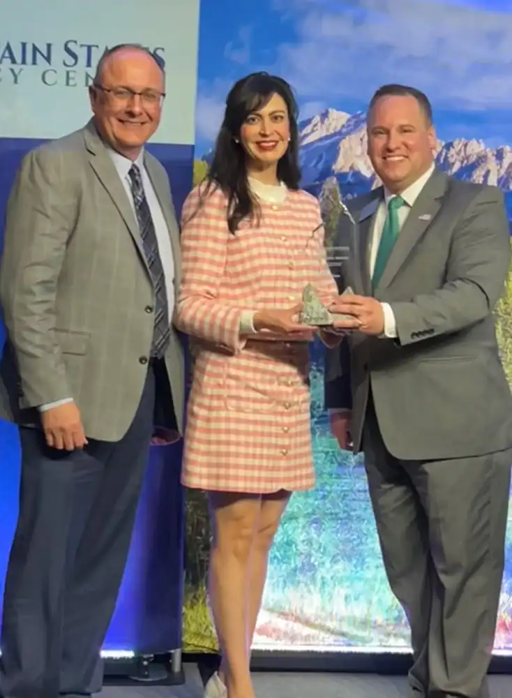 Three people in business attire stand together on a stage. The woman in the center, joined by Secretary Jacobsen and another colleague, holds a glass award and smiles as they pose for a photo against a scenic mountain backdrop.
