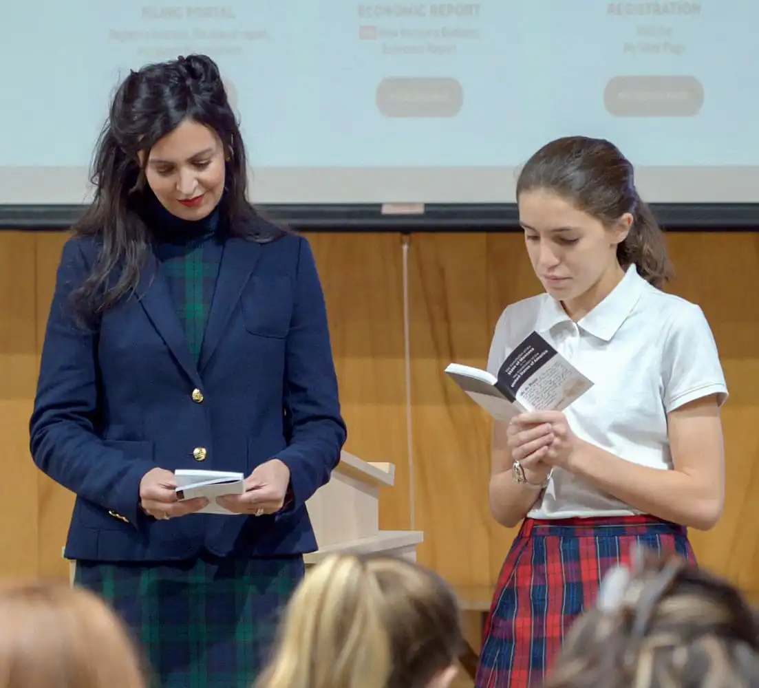 Two women stand at the front of a room. One wears a navy blazer and green plaid skirt, the other a white polo and red plaid skirt. Both are reading from booklets to an audience, with Secretary Jacobsen&rsquo;s name visible on the screen behind them.