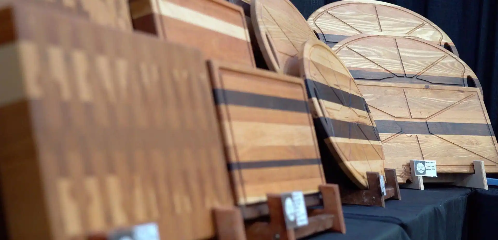 Several wooden cutting boards and serving trays by Montana Block, featuring unique shapes and wood patterns, are displayed upright on a black tablecloth at a craft fair as part of the Made in Montana program.