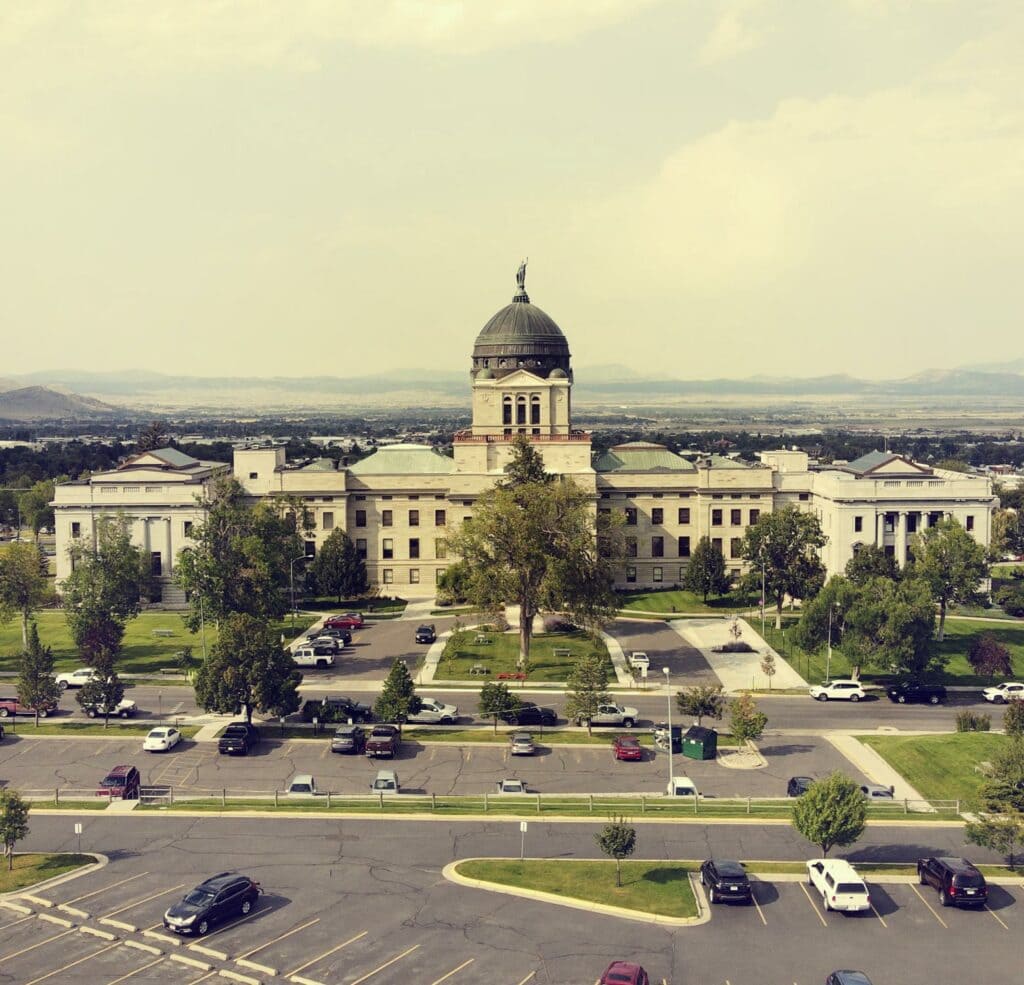 A large government building with a central dome and flag on top, surrounded by trees and lawns, offers business services; parked cars line the front as mountains rise in the background beneath a hazy sky.