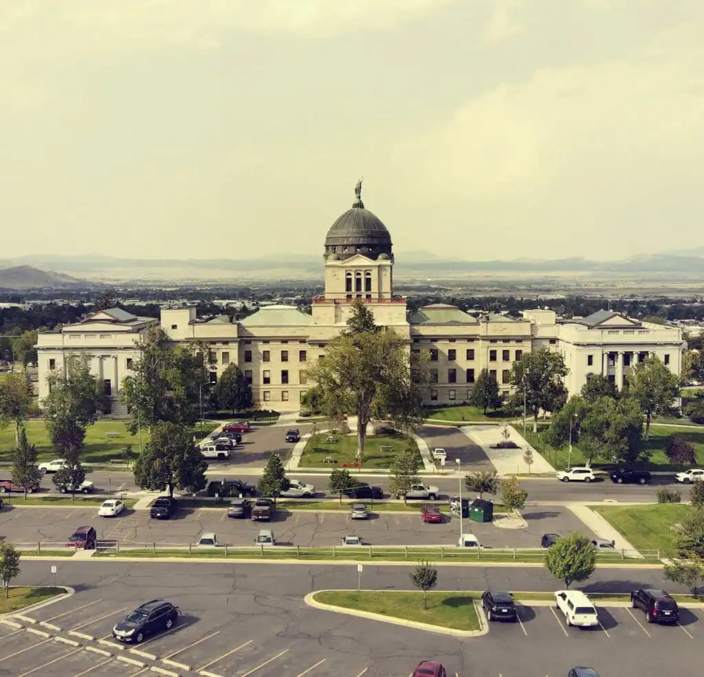 A large government building with a central dome and flag on top, surrounded by trees and lawns, offers business services; parked cars line the front as mountains rise in the background beneath a hazy sky.
