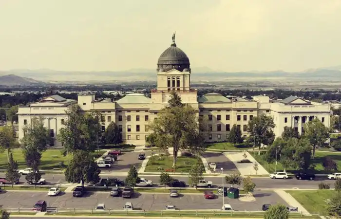 Secretary Christi Jacobsen, state officials to welcome Halloween trick-or-treaters to State Capitol 1 A large government building with a central dome and flag on top, surrounded by trees and lawns, offers business services; parked cars line the front as mountains rise in the background beneath a hazy sky.