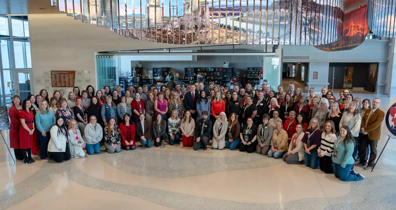 A large group of people, including men and women of various ages, pose for a group photo inside a spacious, modern building with high ceilings, art installations, and natural light alongside Montana Secretary of State Christi Jacobsen.