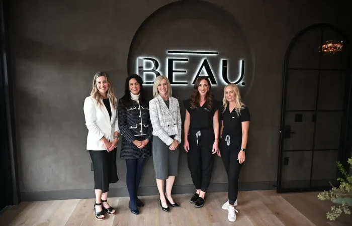 Five women stand smiling in front of a dark wall with a neon sign that reads BEAU, celebrating new business milestones. Dressed in professional and semi-casual attire, they pose together on a light wood floor. Secretary Christi Jacobsen joins the group.
