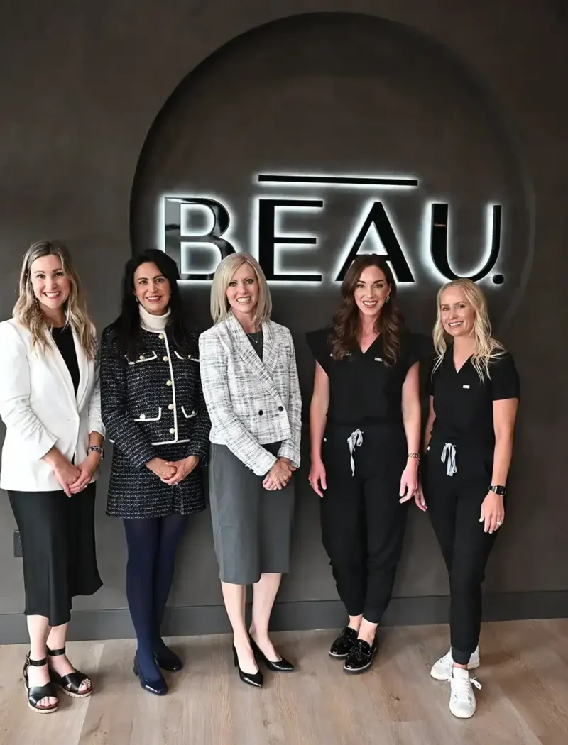 Five women stand smiling in front of a dark wall with a neon sign that reads BEAU, celebrating new business milestones. Dressed in professional and semi-casual attire, they pose together on a light wood floor. Secretary Christi Jacobsen joins the group.