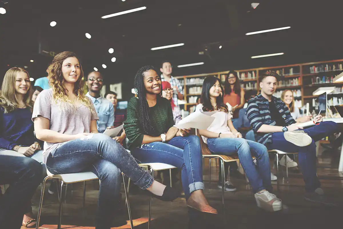 A diverse group of students sits on chairs in a library or classroom, smiling and looking forward, some holding papers and notebooks related to Civics. Bookshelves are visible in the background, and the YouthVote atmosphere appears positive and engaged.