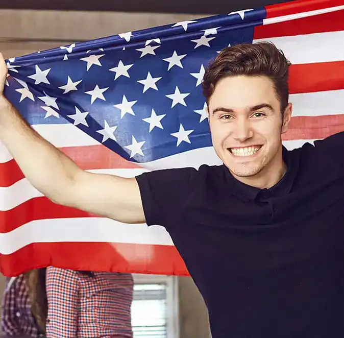 A smiling young man in a black shirt holds up an American flag behind him, stretching it wide with both arms—capturing the spirit of YouthVote and pride in civics.
