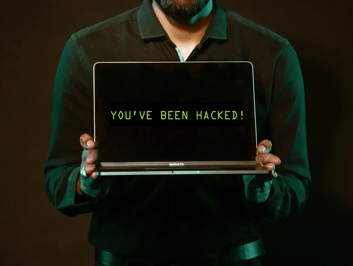 A person in a dark shirt holds a laptop, with the screen displaying the message YOUVE BEEN HACKED! in green text on a black background.
