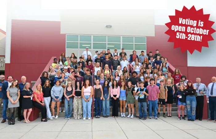 A large group of students and adults stand on school steps smiling. A red banner says, Voting is open October 5th-28th! The building is red and white with large windows, highlighting the excitement around YouthVote and promoting civic engagement among the community.