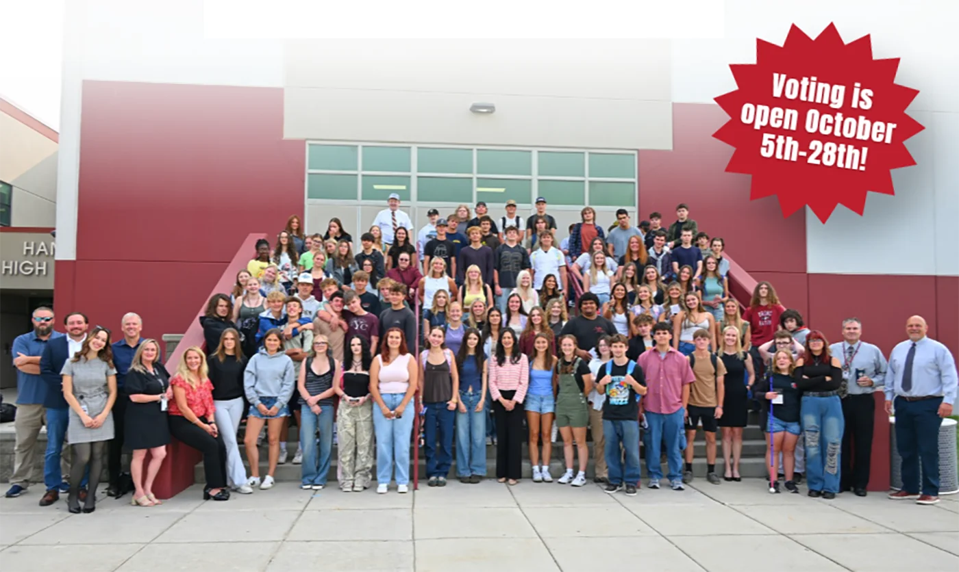 A large group of students and adults stand on school steps smiling. A red banner says, Voting is open October 5th-28th! The building is red and white with large windows, highlighting the excitement around YouthVote and promoting civic engagement among the community.