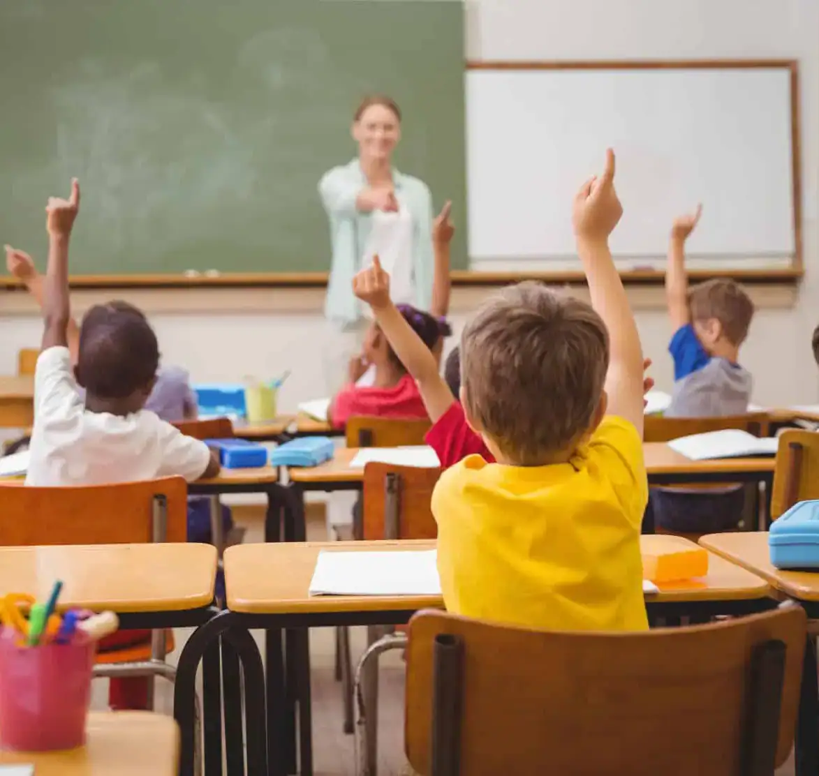 Children sit at desks in a classroom, raising their hands while a teacher stands at the front near a chalkboard, engaging with the students in a lively civics lesson. School supplies are visible on the desks.
